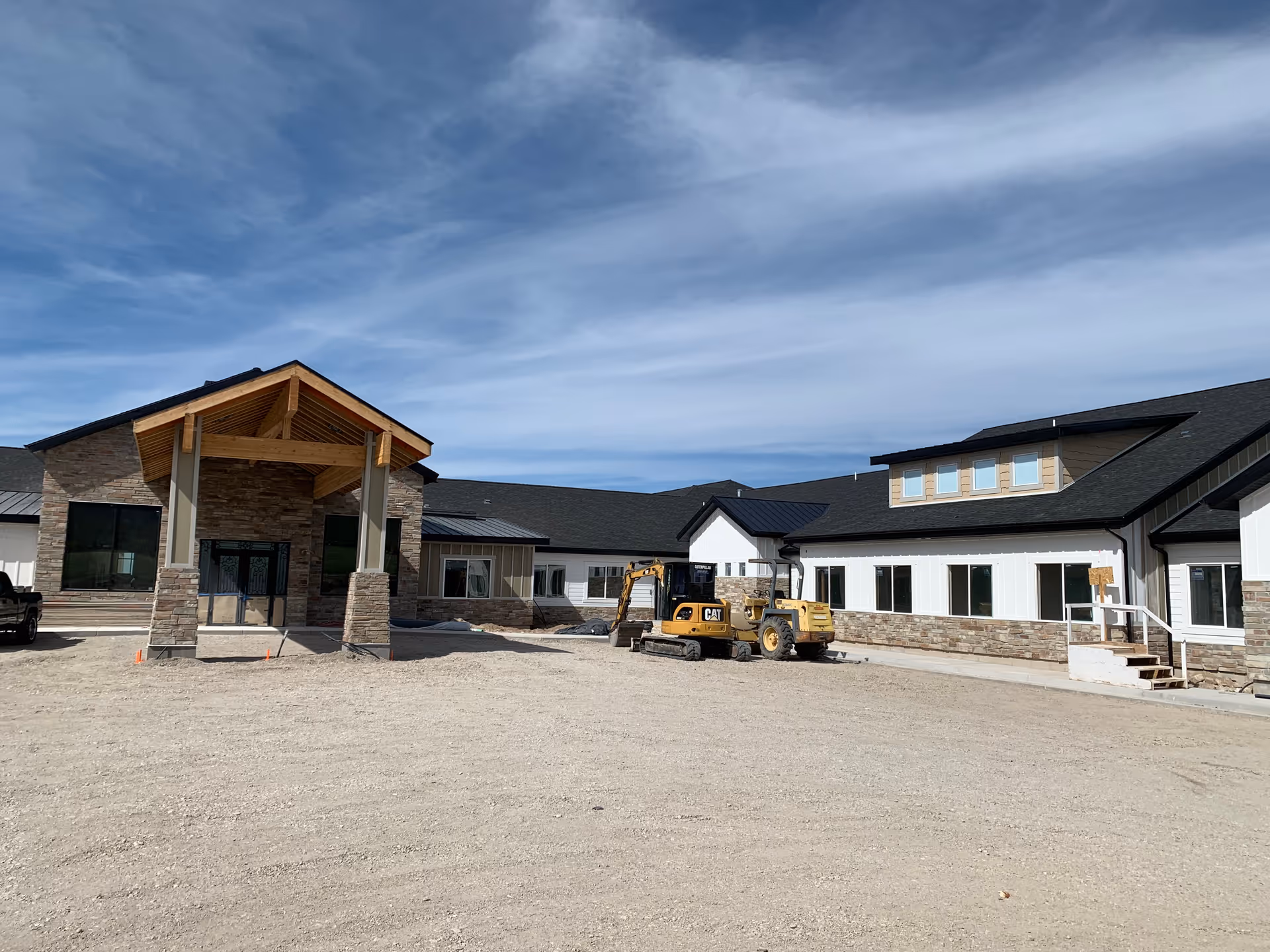 Exterior view of a newly constructed assisted living facility building with stone and white siding, a covered entrance with wooden beams, and construction equipment parked in front on a dirt lot under a partly cloudy sky.