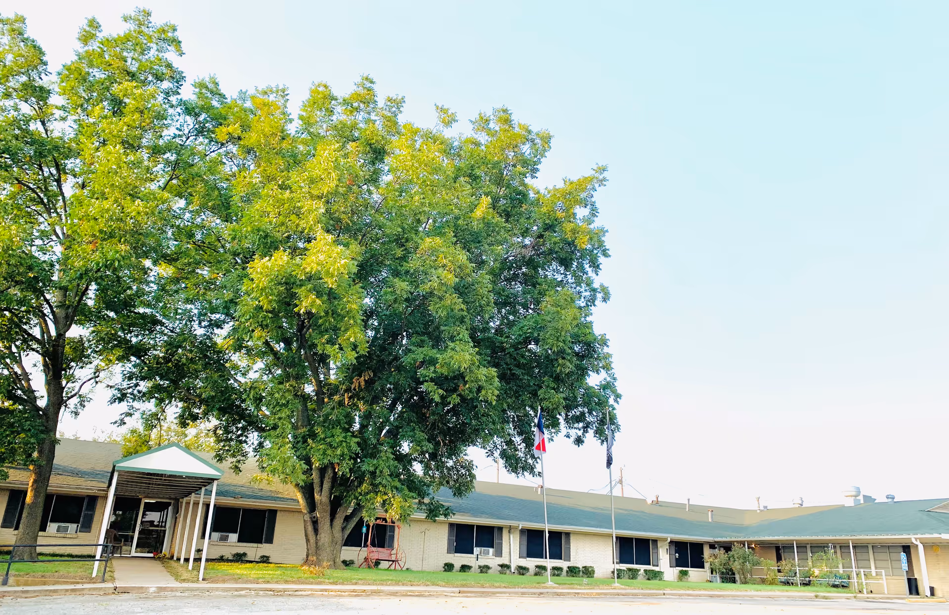 Exterior view of Western Hills Healthcare Residence showing a single-story building with a green roof, large trees in front, a covered entrance, and two flagpoles with flags.