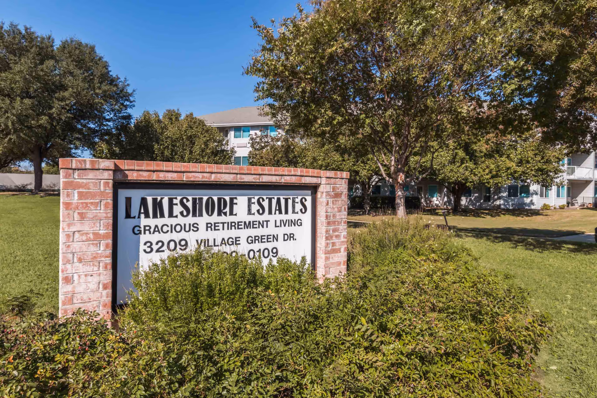 Brick entrance sign reading 'Lakeshore Estates Gracious Retirement Living' in front of a multi-story residential building with trees and lawn.