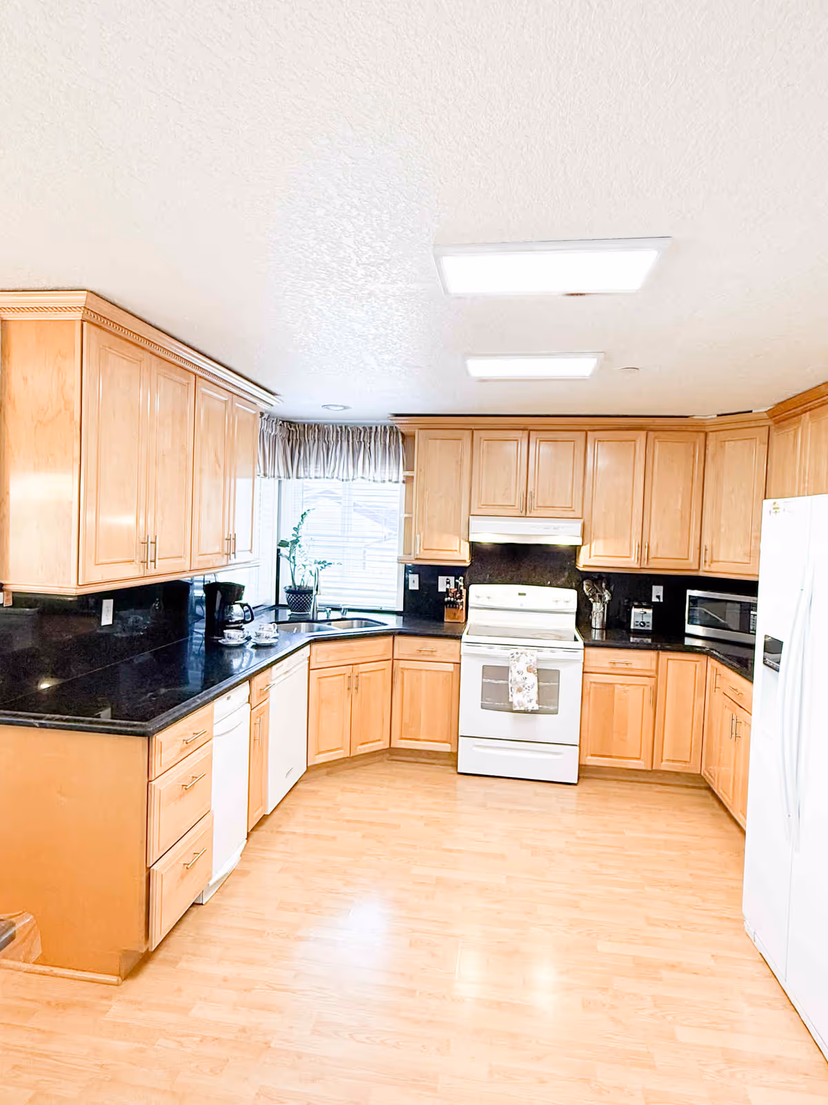 Bright U-shaped kitchen with light wood cabinets, black countertops, and white appliances.