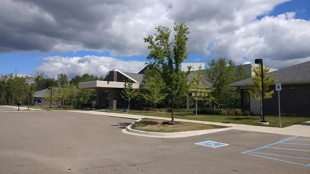 Exterior view of WellBridge of Grand Blanc showing the facility entrance, parking area with handicap spaces, and landscaped trees under a cloudy sky.