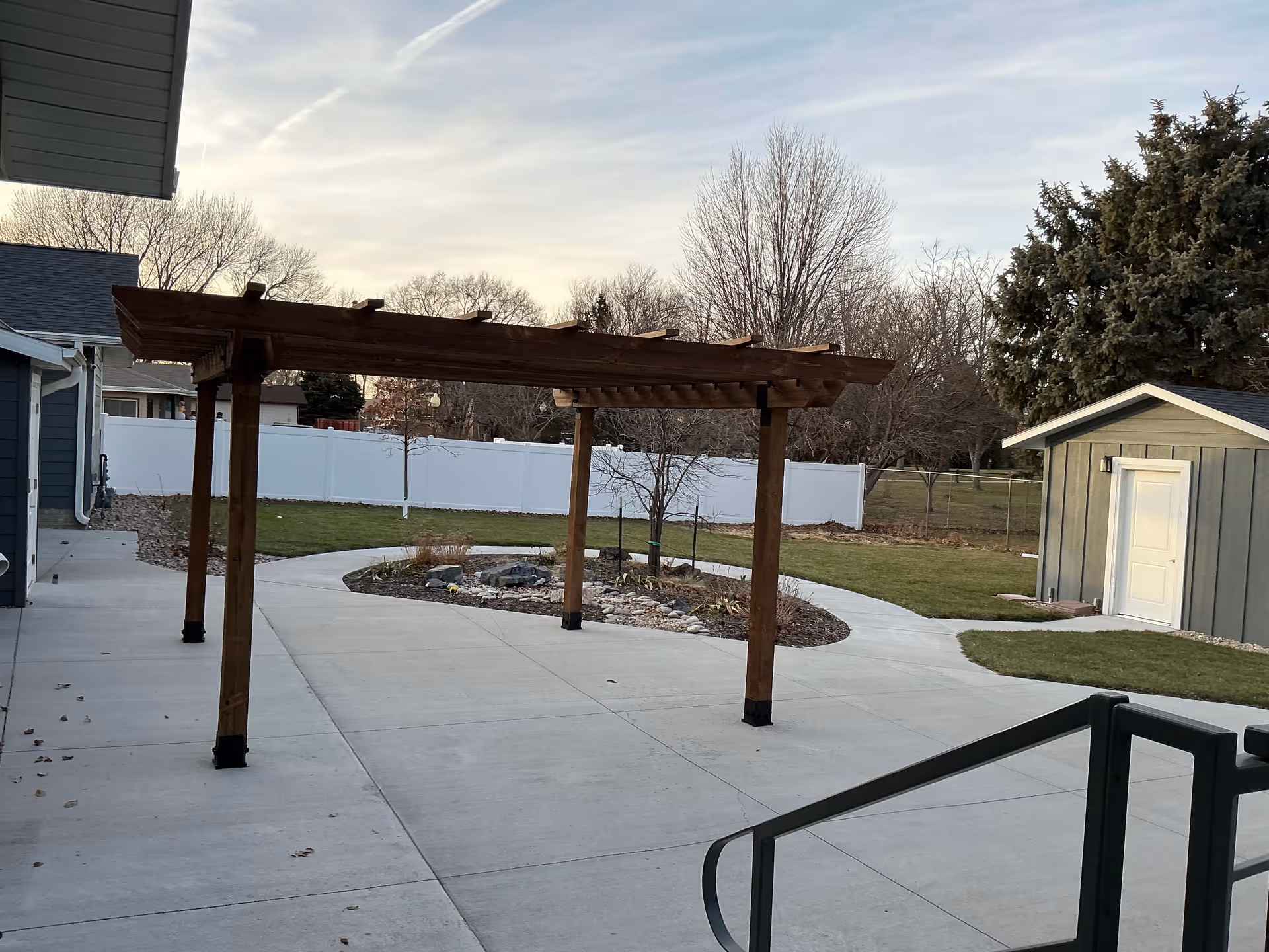 Exterior courtyard featuring a wooden pergola, concrete pathways, garden bed, shed, and fence.