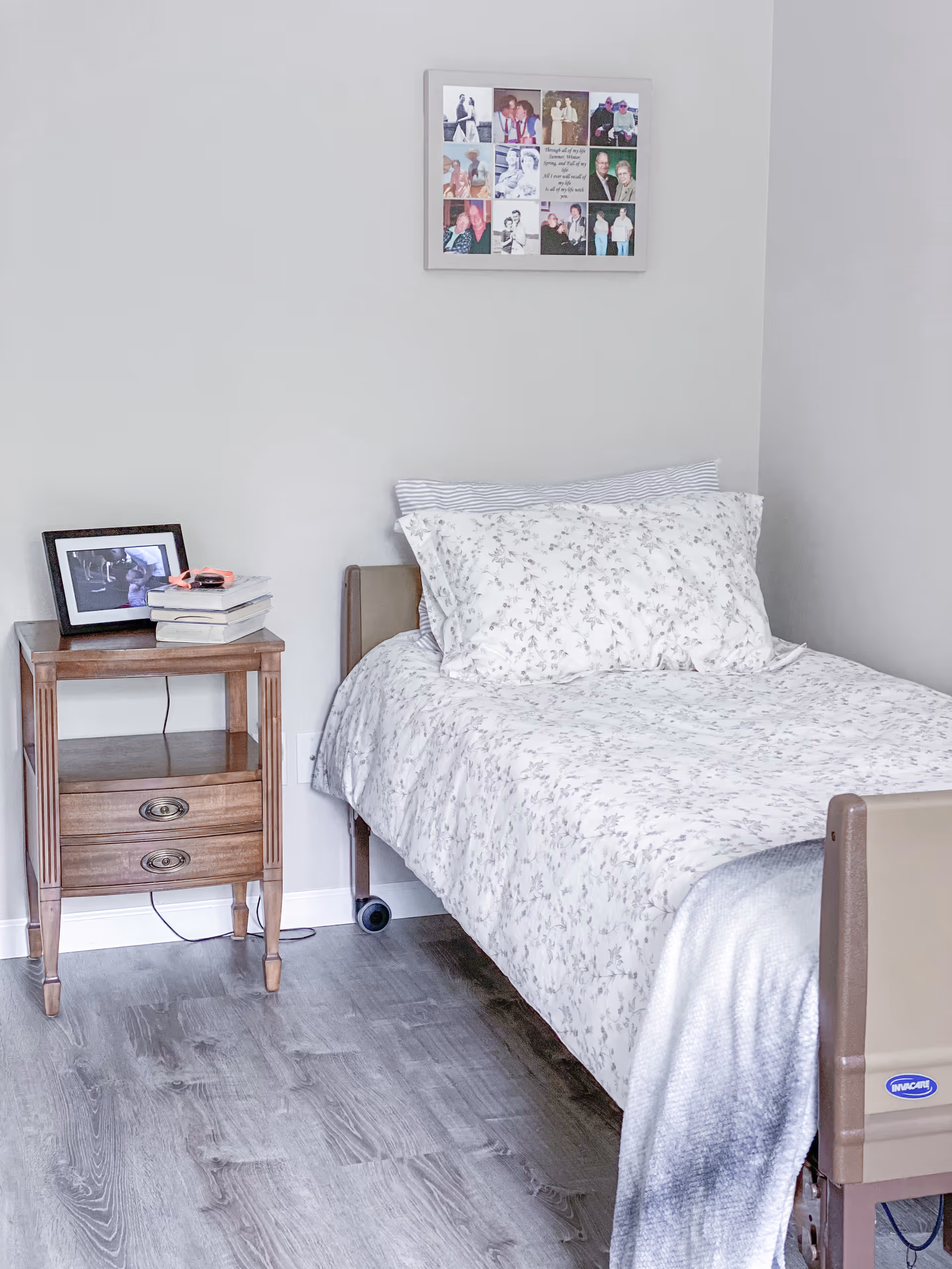 A tidy bedroom in a senior living facility with a bed, bedside table, and framed photos.