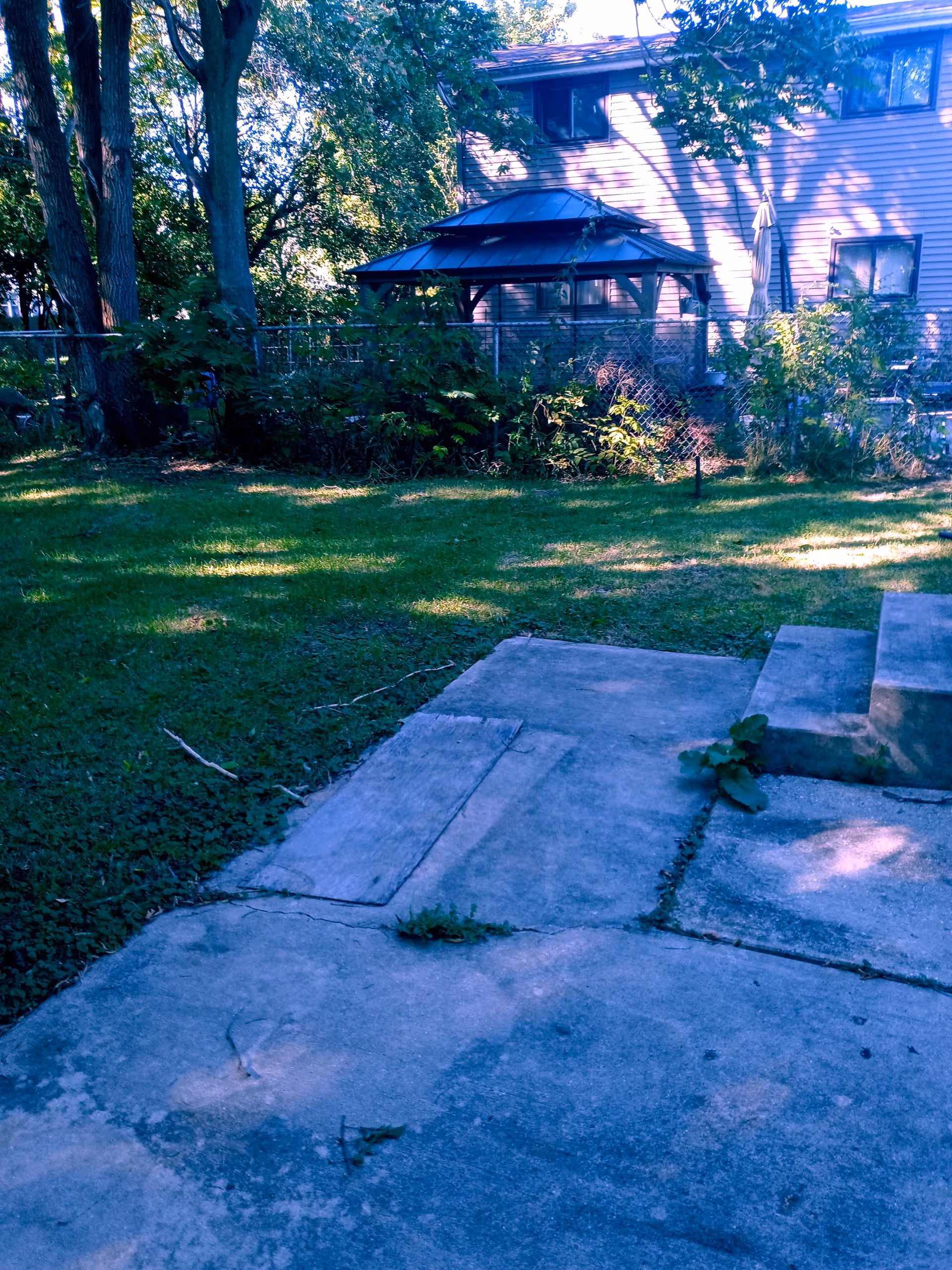 Backyard view of concrete patio, steps, lawn, fence, and gazebo behind residential building.