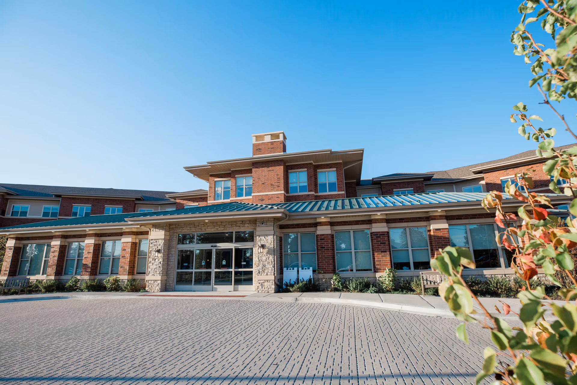 Front exterior view of a senior living facility building with brick and stone facade, large windows, and a green metal roof under a clear blue sky. There are some plants and bushes near the entrance and a paved driveway in front.