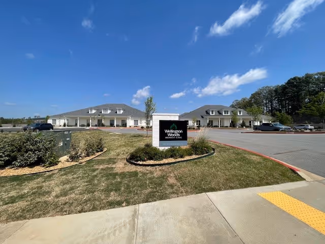 Front exterior of Wellington Woods Memory Care with a lawn, entrance sign, and two single-story buildings under a blue sky.