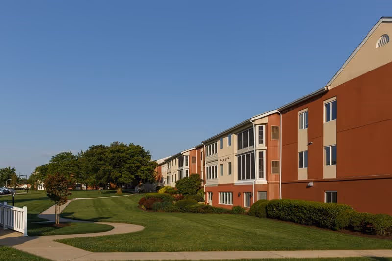 Exterior view of a multi-story senior living building with a manicured lawn, walkway, and trees under a clear blue sky.