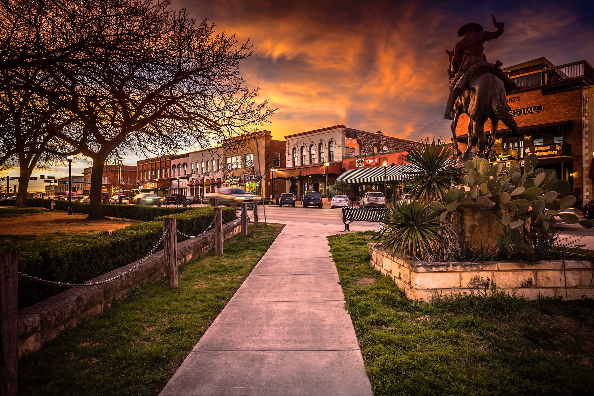 A scenic view of a small town street at sunset with a sidewalk leading through a park area. On the right side, there is a statue of a person on horseback surrounded by plants. Buildings with storefronts and parked cars line the street under a dramatic orange and purple sky.