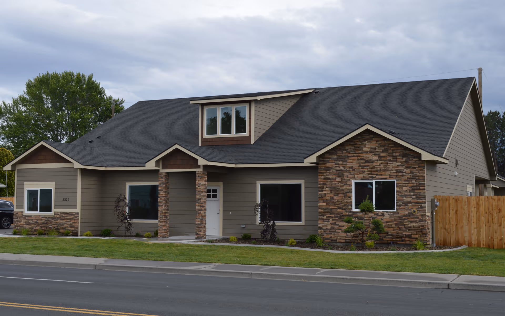 Single-story residential building with gray siding and stone accents, a front lawn and sidewalk under a cloudy sky.