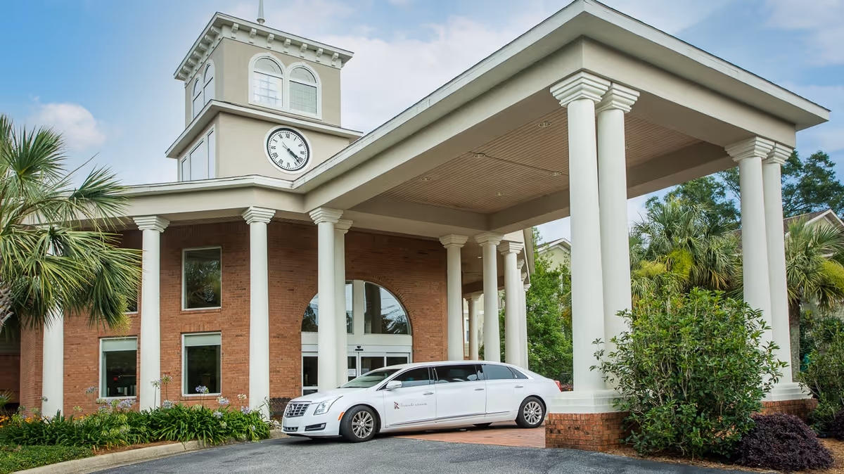 Front exterior view of a senior living facility named Veranda of Pensacola, featuring a brick building with large white columns supporting a covered entrance. A white limousine is parked under the entrance canopy. The building has a clock tower and is surrounded by palm trees and greenery under a blue sky with some clouds.