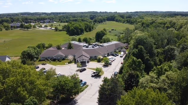 Aerial view of Watson Fields senior living facility surrounded by lush green trees and open fields under a clear sky. The building has a large parking area with several cars parked, and the facility is situated in a peaceful, rural setting.