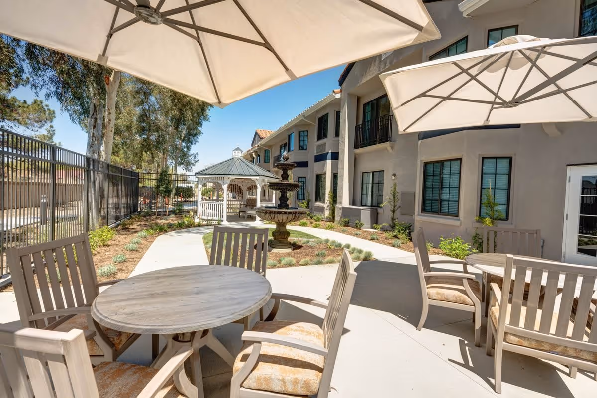Outdoor patio area at The Havens at Antelope Valley featuring round tables with cushioned chairs under large umbrellas, a paved walkway, landscaped garden beds, a multi-tiered fountain, and a white gazebo next to a two-story building under a clear blue sky.