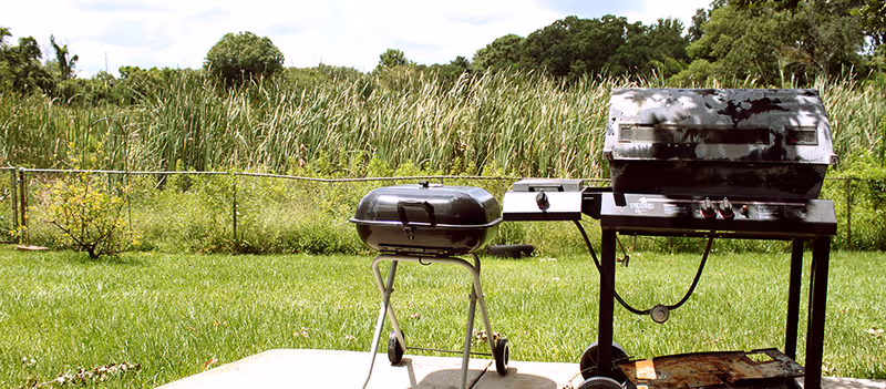 Two outdoor grills on a concrete patio with green grass, a chain-link fence, and tall reeds or grasses in the background under a partly cloudy sky.