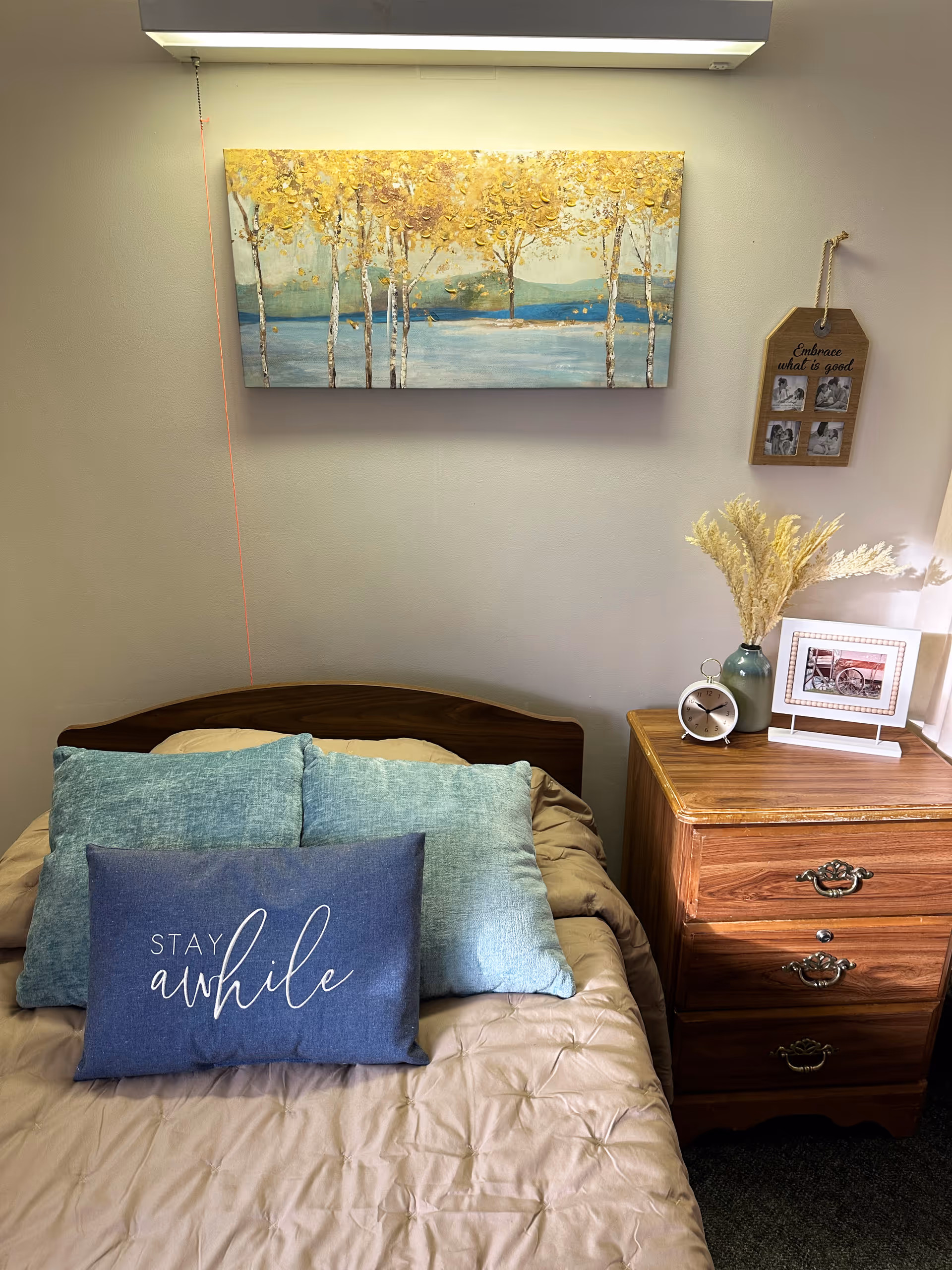 A cozy bedroom setup with a bed featuring three pillows, one of which has the text 'Stay awhile'. Above the bed is a painting of trees with yellow leaves and a lake in the background. To the right of the bed is a wooden nightstand with a small clock, a vase with dried flowers, and a framed photo. A small hanging plaque on the wall reads 'Embrace what is good' with four small photos.