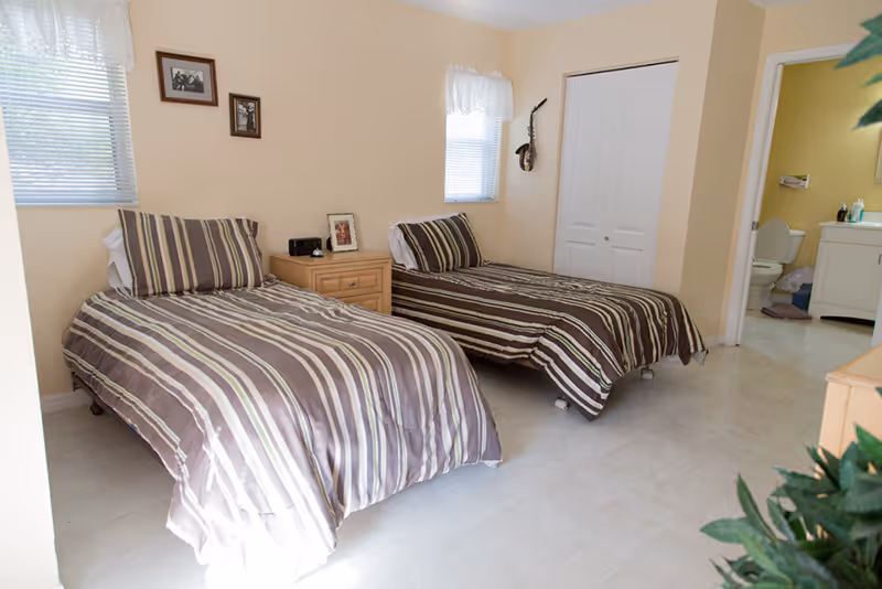 Two twin beds with striped bedding, a nightstand, and windows in a beige senior living bedroom.