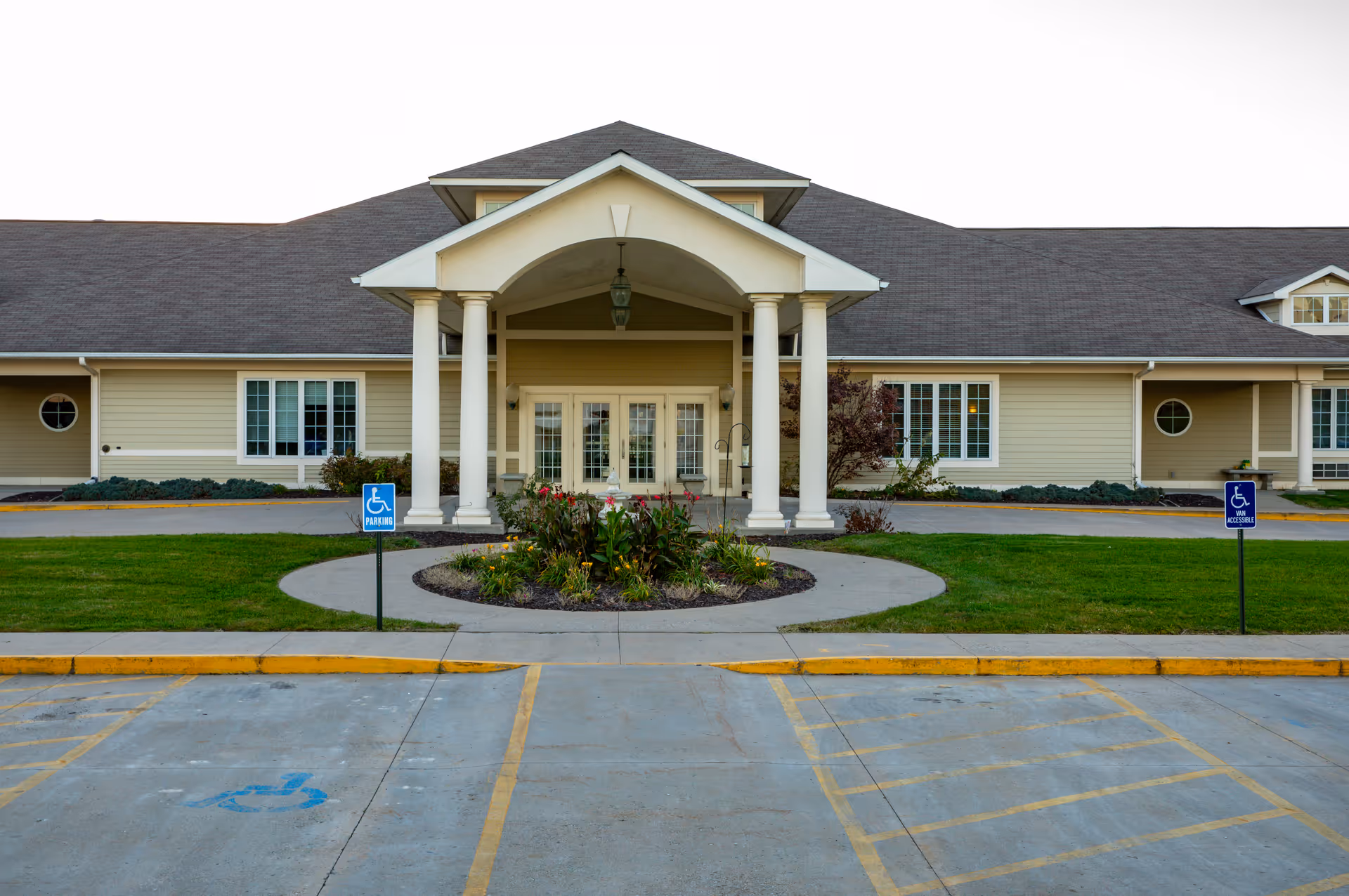 Front exterior view of Homestead Assisted Living & Memory Care of Centerville building with a covered entrance supported by four white columns, a circular flower bed with plants in front, and two handicap parking signs on either side of the entrance. The building has beige siding, white trim, and multiple windows.