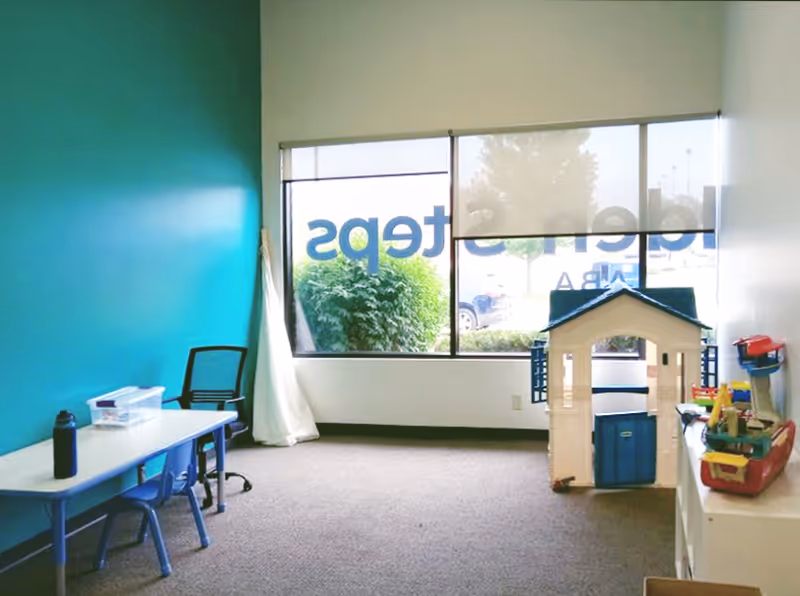 Bright therapy playroom with teal wall, large window, toy house, and table.