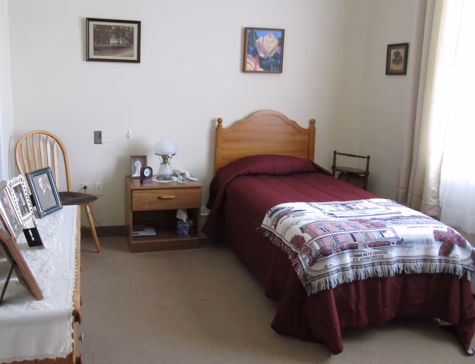 A simple assisted-living bedroom with a single bed dressed in burgundy bedding, a nightstand with a lamp, a dresser with framed photos, and a wooden chair.