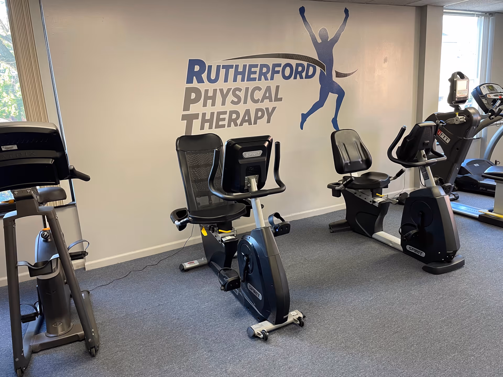 Indoor physical therapy gym featuring exercise bikes and a Rutherford Physical Therapy mural.