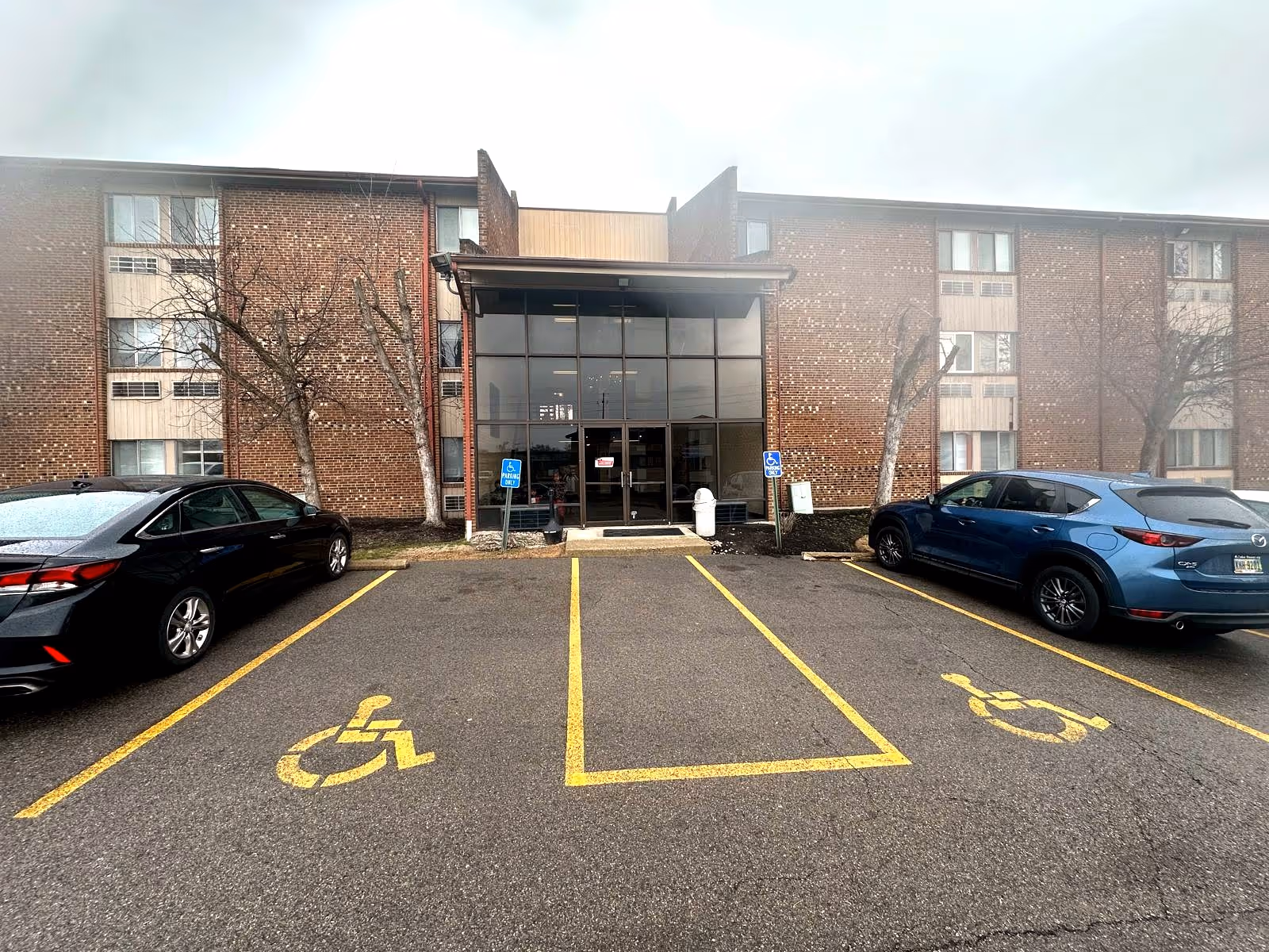 Brick senior living facility entrance with glass doors and parked cars in the lot.