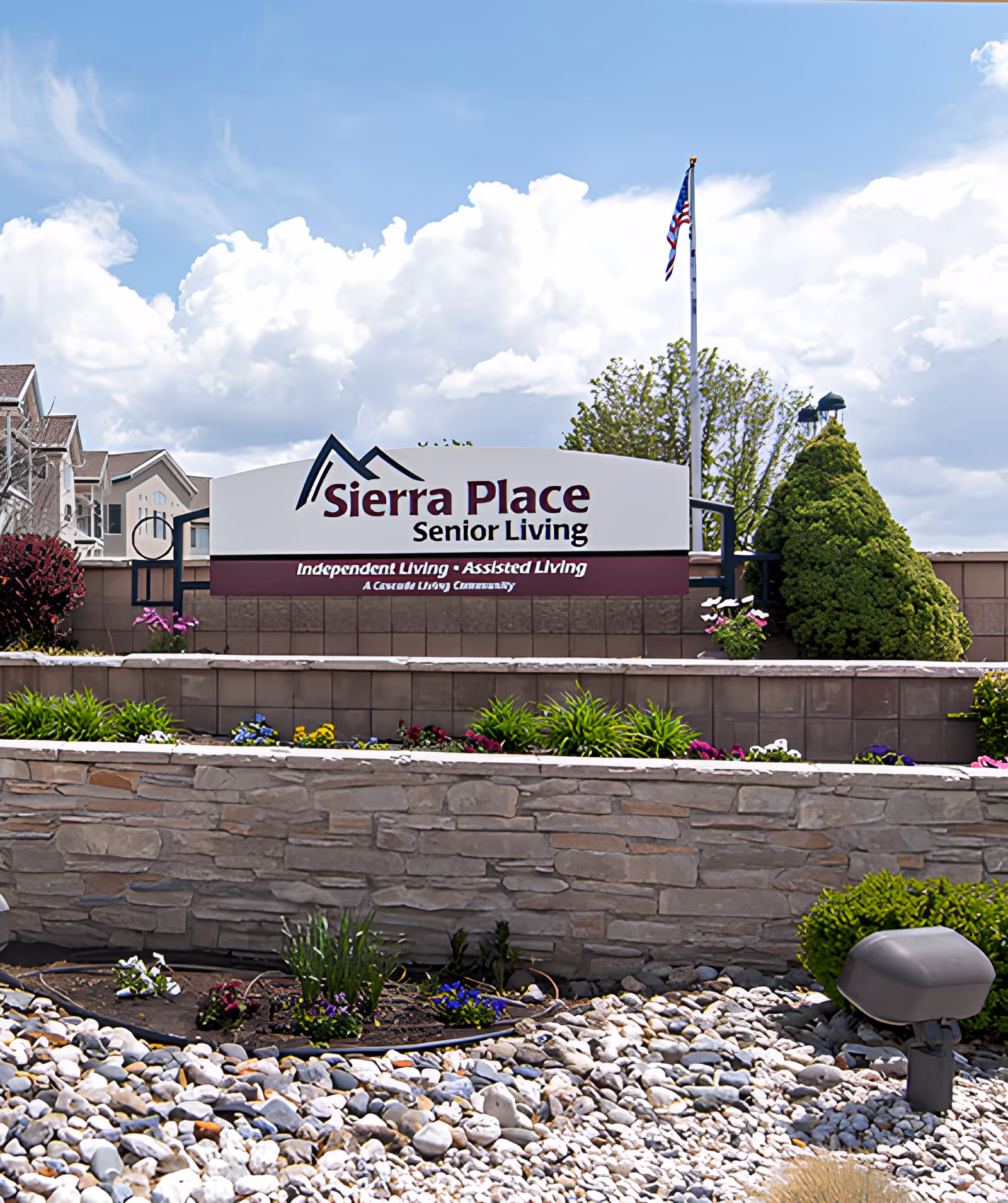 Outdoor view of the entrance sign for Sierra Place Senior Living, featuring a landscaped area with flowers, bushes, and rocks in front of a stone wall. The sign displays the facility name and mentions Independent Living and Assisted Living. An American flag is visible on a flagpole behind the sign under a partly cloudy sky.