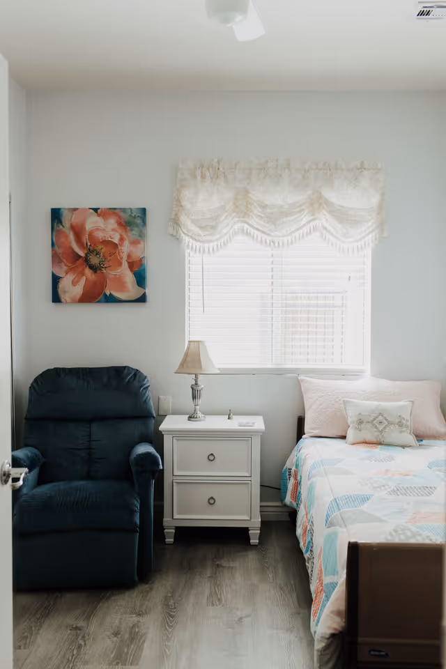 Cozy bedroom with a bed, nightstand, lamp, chair, window, lace valance, and floral art.