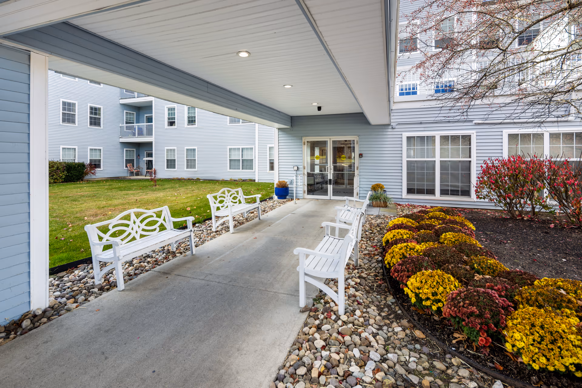 Front entrance of a blue senior living building with white benches along a rocky path.