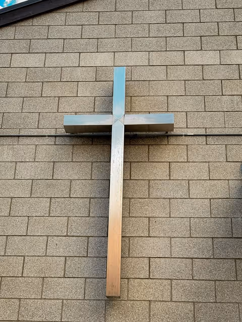Exterior view of a metallic cross mounted on a beige brick church wall.