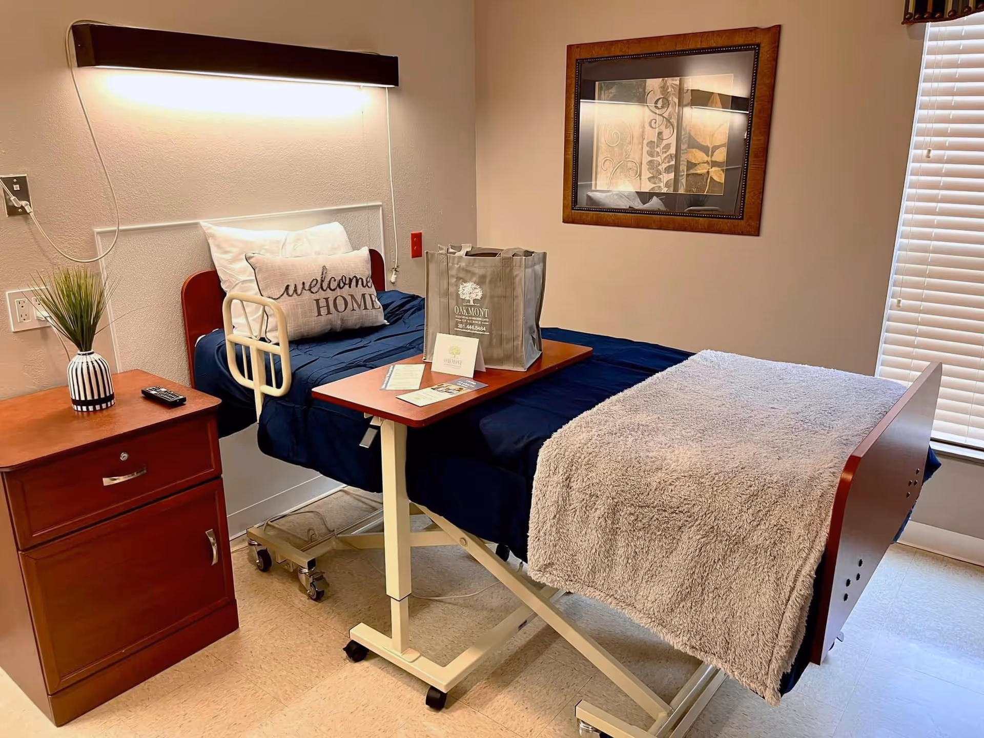 A neatly arranged healthcare facility bedroom with a hospital bed covered in dark blue bedding and a light gray blanket. There is a 'welcome HOME' pillow on the bed. A wooden bedside table with a small plant and a remote control is next to the bed. A wooden overbed table holds a branded Oakmont Healthcare & Rehabilitation Center tote bag and some brochures. A framed artwork hangs on the wall, and a window with blinds is partially visible.