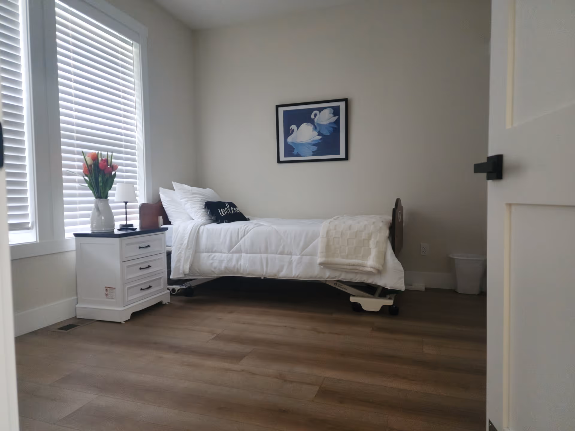 A tidy bedroom with a bed, white bedding, nightstand, blinds, and wall art.