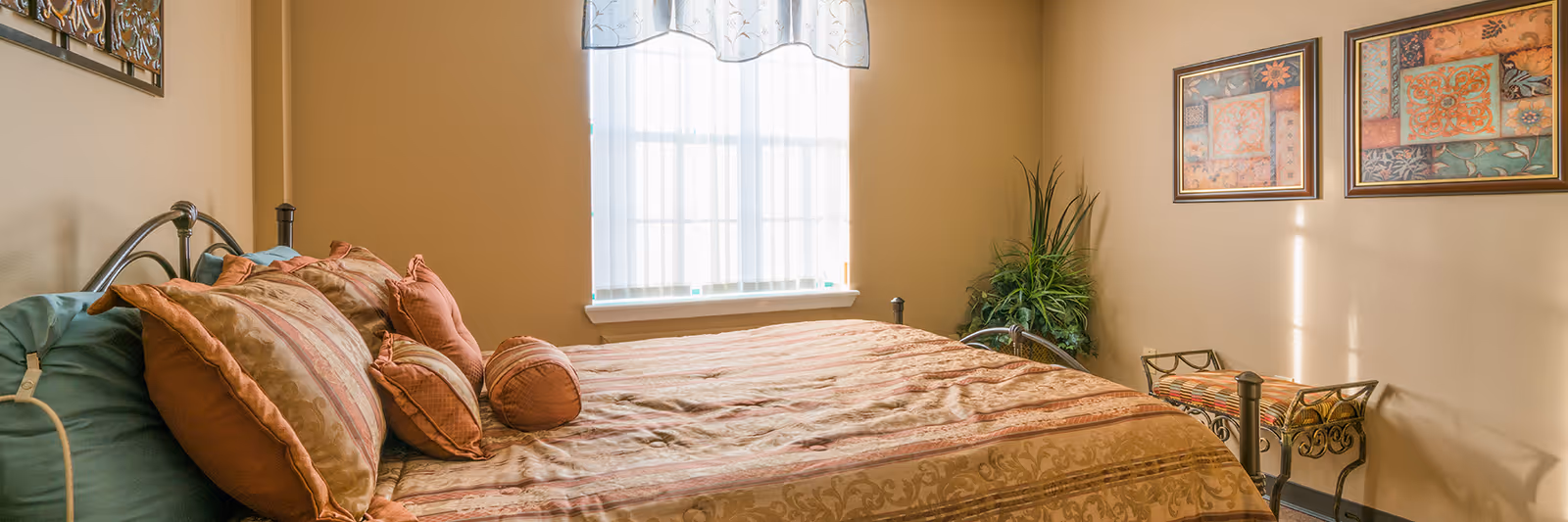 A cozy bedroom with a large bed covered in a patterned bedspread and multiple pillows. The room has beige walls, a window with sheer curtains letting in natural light, two framed artworks on the wall, a decorative bench with a colorful cushion, and a green potted plant in the corner.