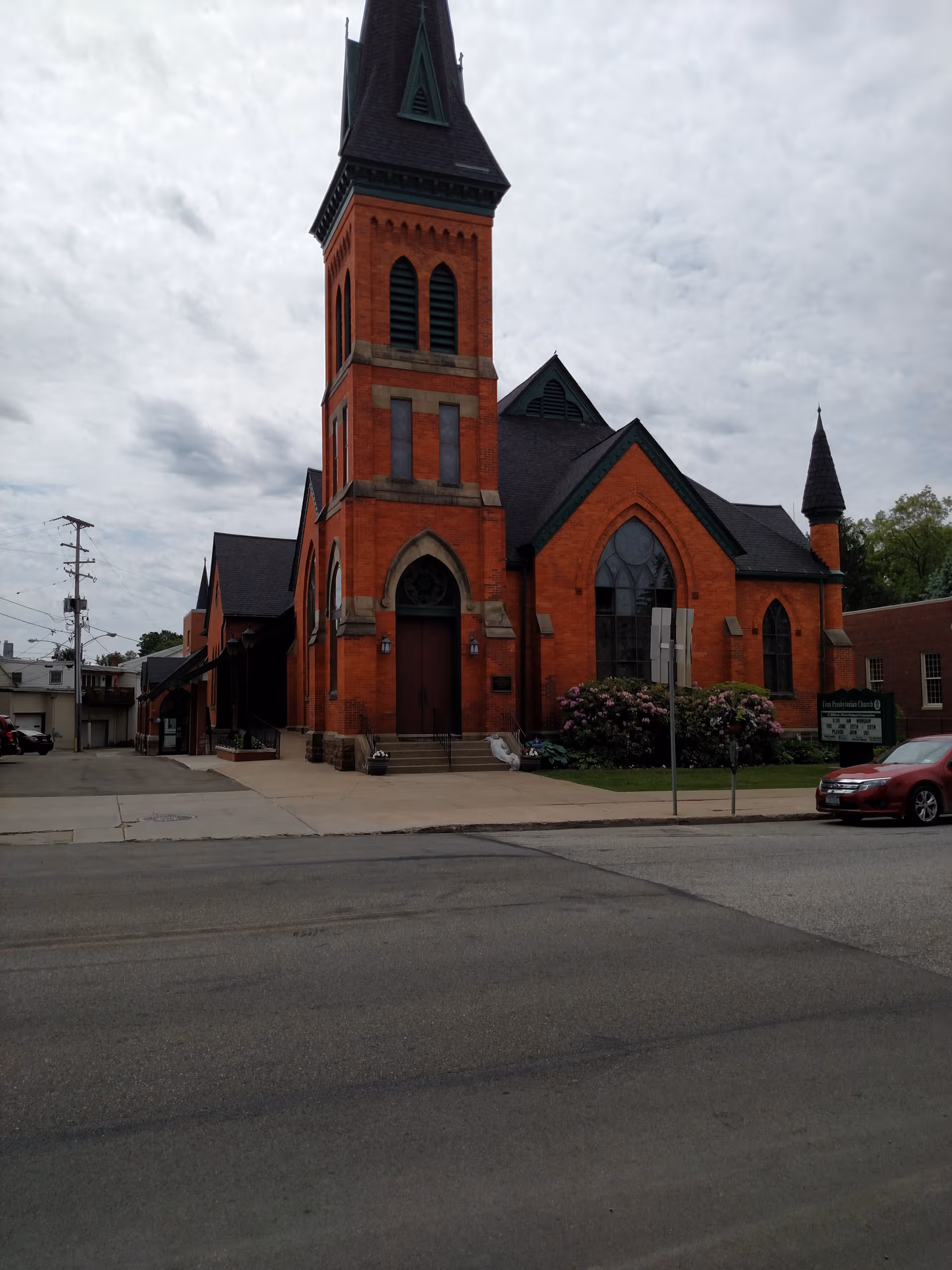 Red brick church with tall steeple, arched windows, and a sidewalk along a street