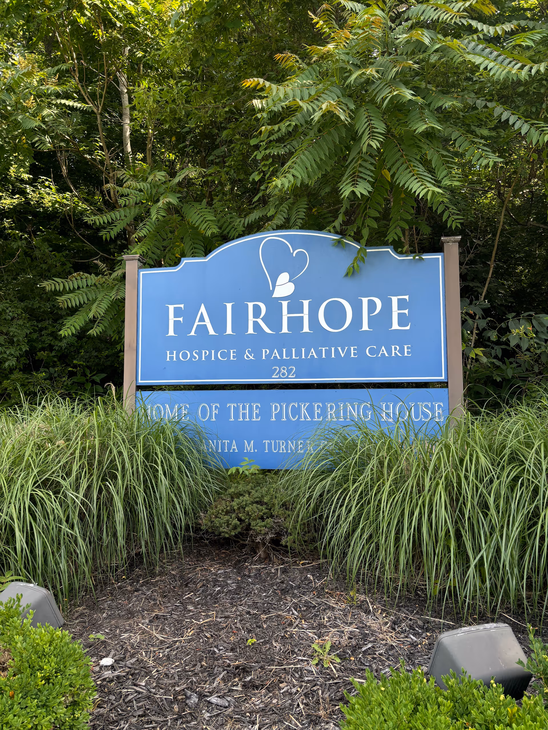 Blue FAIRHOPE Hospice & Palliative Care sign amid greenery and plants.