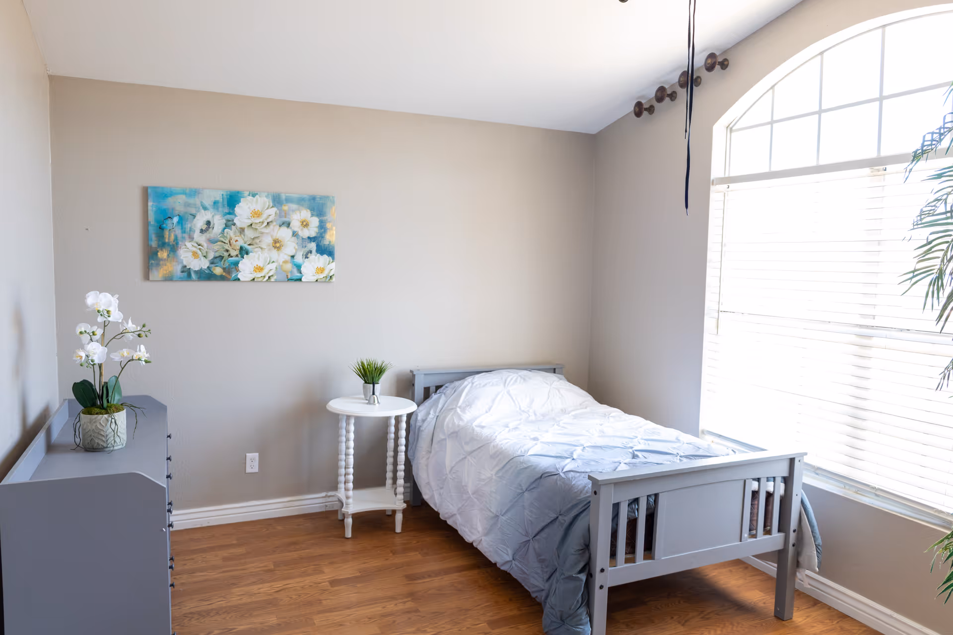 Small, neatly arranged bedroom with single bed, nightstand, dresser, and large sunlit window.
