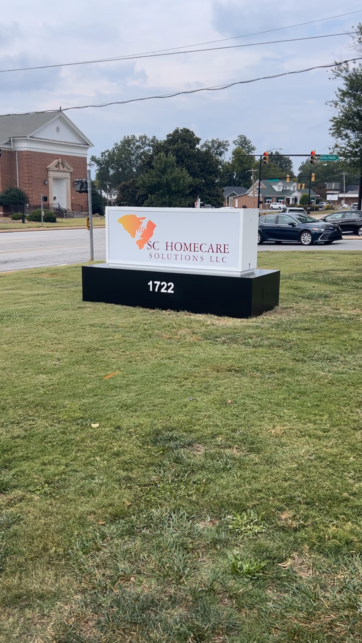 Outdoor sign on a grassy lot for SC HomeCare Solutions LLC, with street traffic in background.