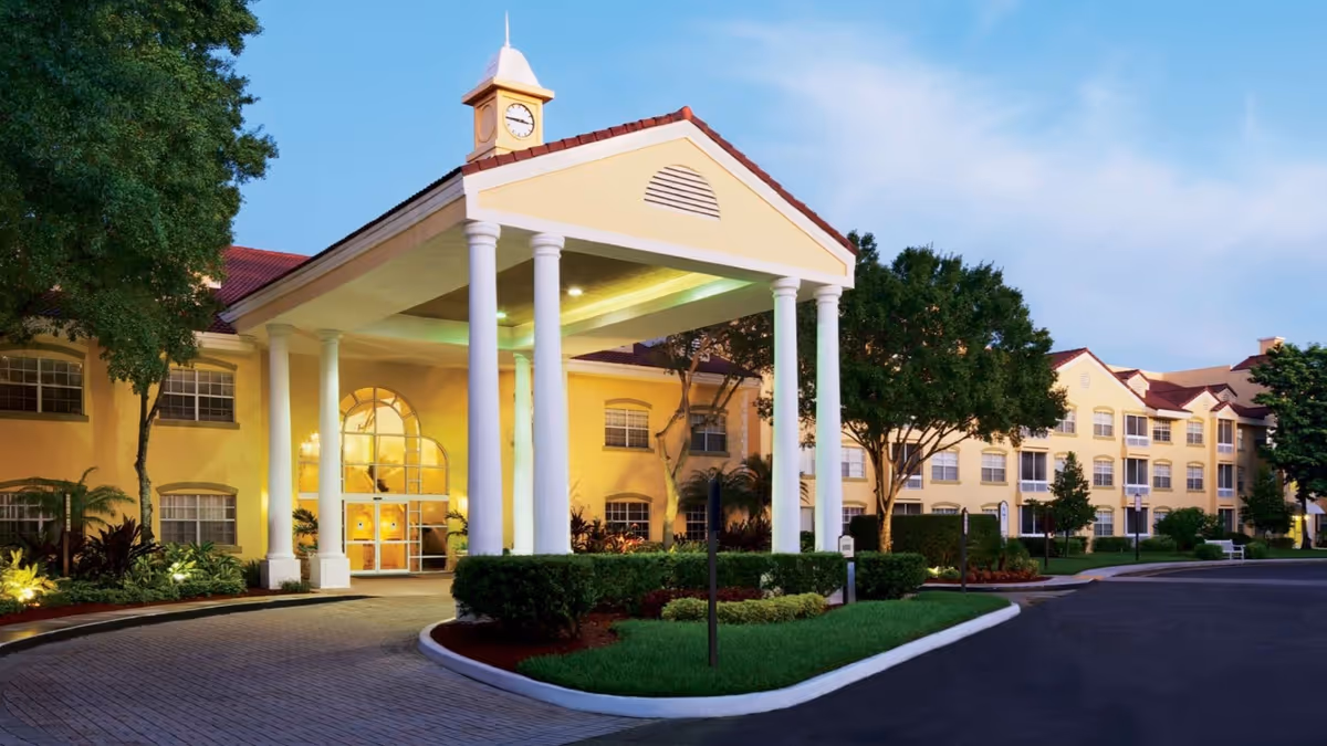 Front view of a beige senior living building with a colonnaded entrance and manicured landscaping.