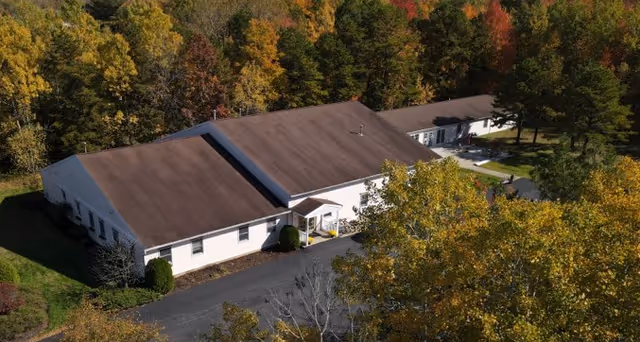 A single-story white building with a dark brown roof, surrounded by autumn trees.