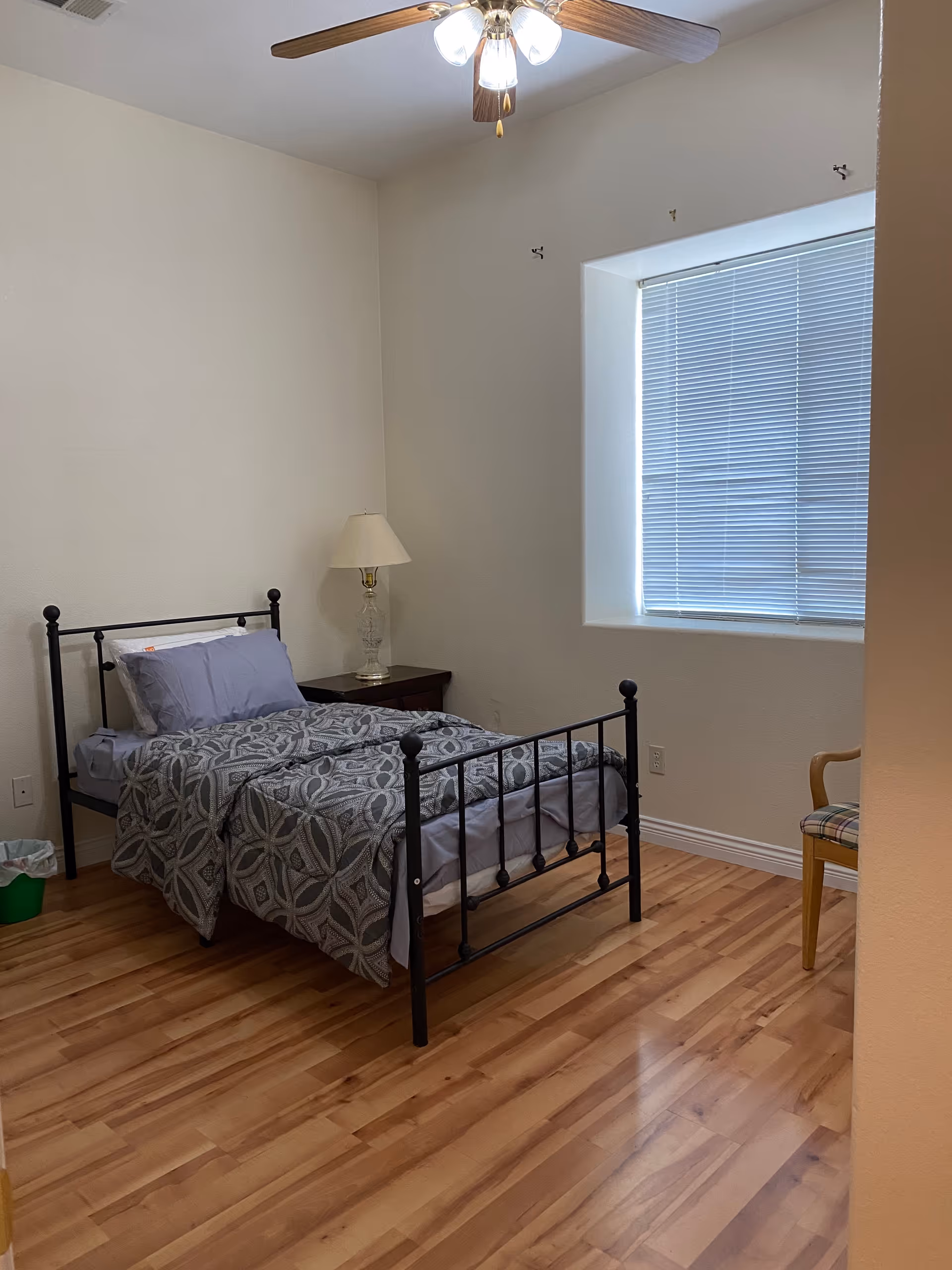 A simple bedroom with a metal bed, nightstand, lamp, blinds, and wooden floor.