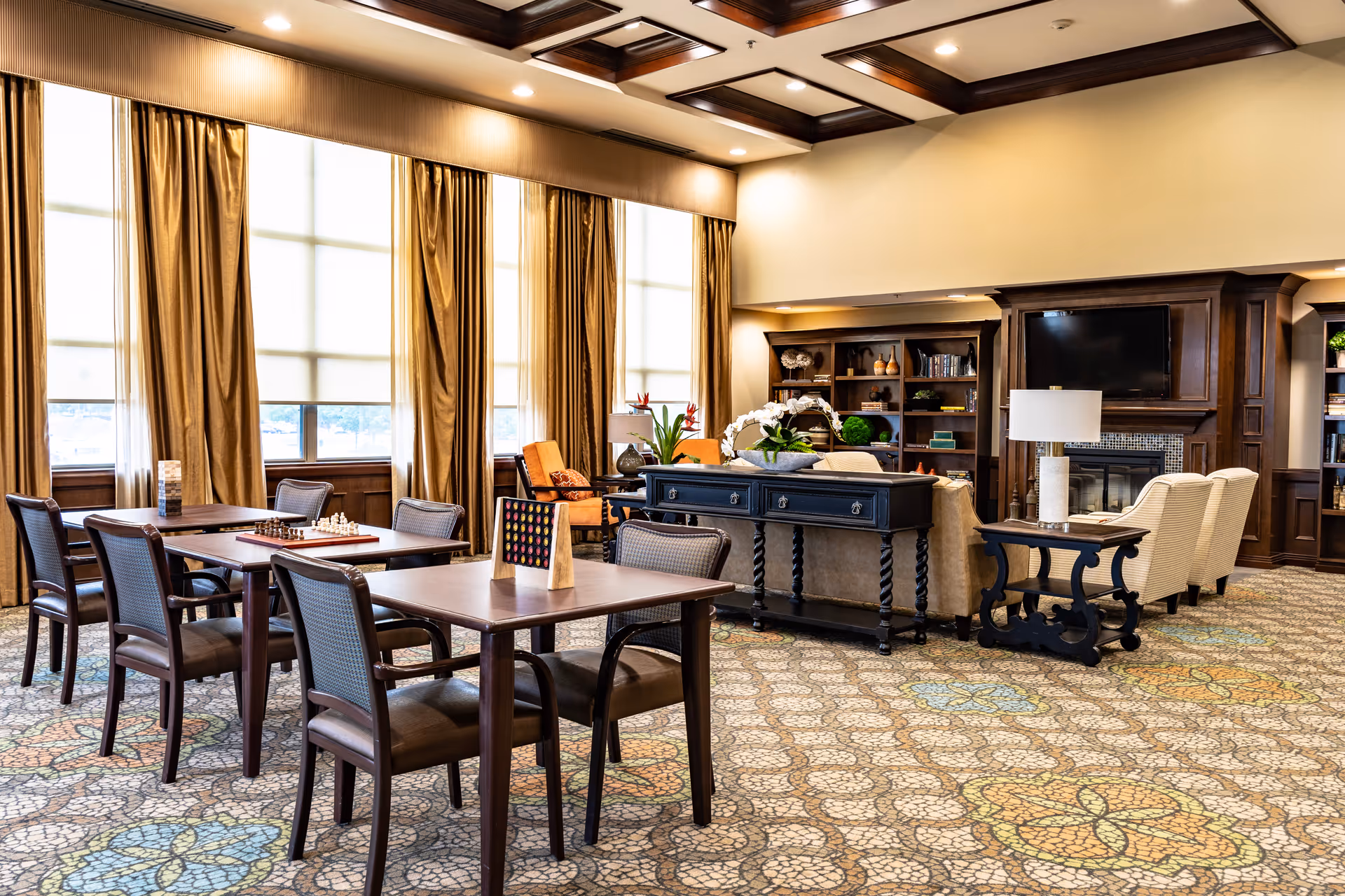 Spacious senior living facility lounge with tables, chairs, a sofa, fireplace, and bookshelves.