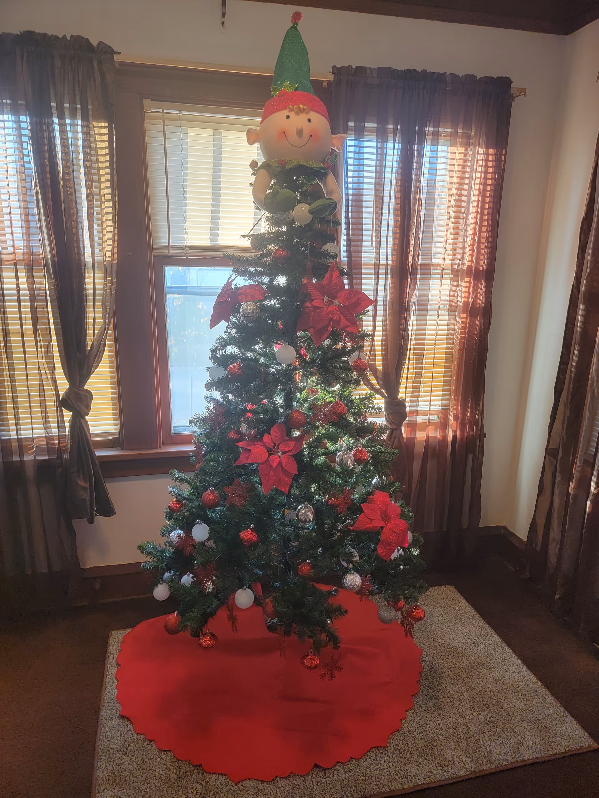 Decorated Christmas tree with red bows and a cheerful elf topper in a sunlit living room.