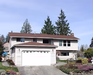 Two-story light-colored house with an attached two-car garage, driveway, landscaped front yard, and tall evergreen trees behind it.