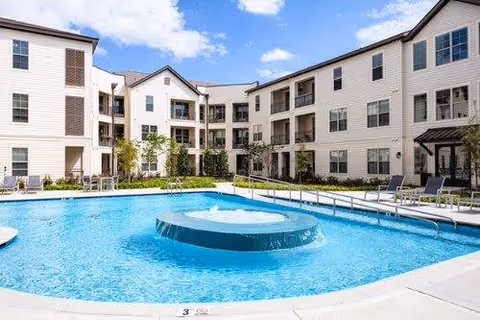 Outdoor swimming pool with a circular water feature in the center, surrounded by lounge chairs and a three-story residential building with balconies in the background under a partly cloudy sky.