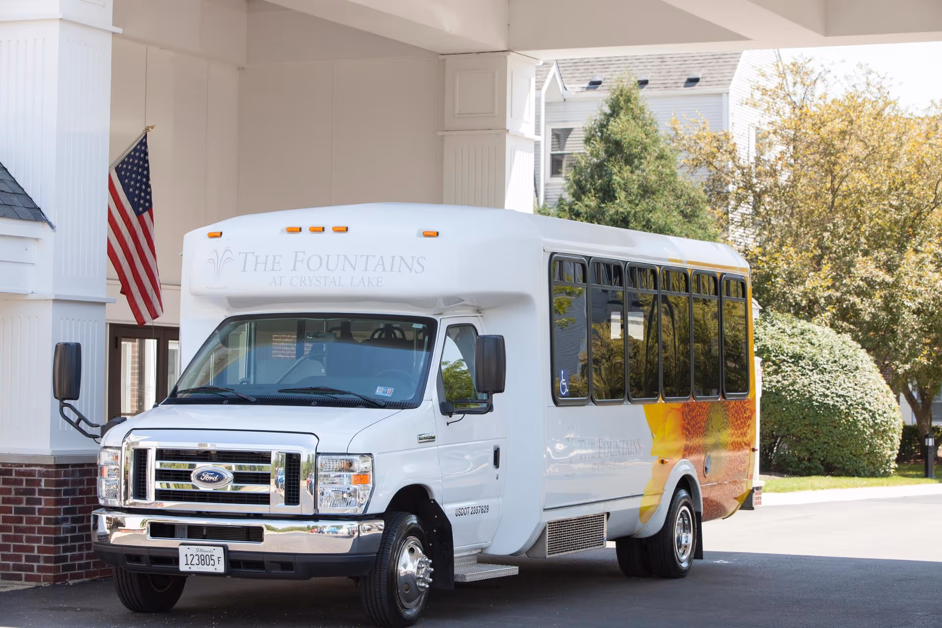 A white shuttle bus with 'The Fountains at Crystal Lake' written on the front is parked under a covered entrance of a building. An American flag is mounted on a white column to the left, and trees and bushes are visible in the background.