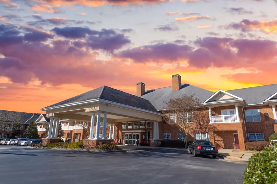 Exterior view of a senior living facility building with a covered entrance and columns, parked cars, and a colorful sunset sky with clouds in the background.