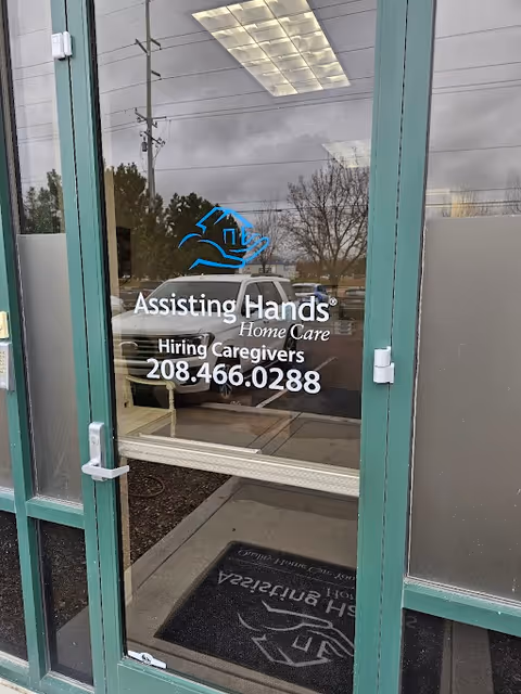 Glass entrance door displays Assisting Hands Home Care branding, hiring caregivers, and phone number.