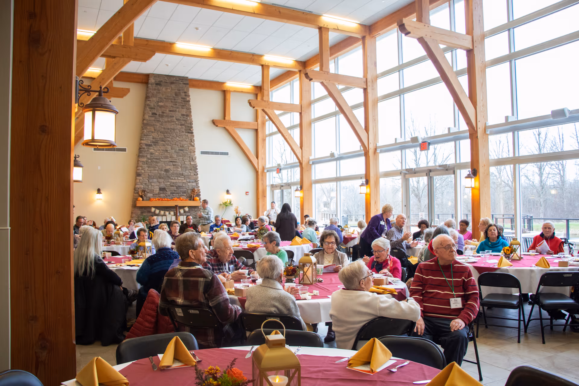 A large dining room filled with elderly people seated at round tables covered with red tablecloths and yellow napkins. The room features tall wooden beams, large windows letting in natural light, and a stone fireplace in the background. The atmosphere appears social and lively.