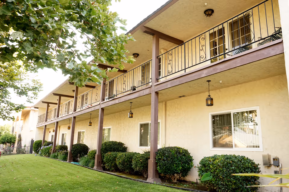 Exterior view of a two-story senior living facility building with beige walls and brown wooden railings on the upper balcony. The building is surrounded by green bushes and a well-maintained lawn, with a large tree providing shade on the left side.