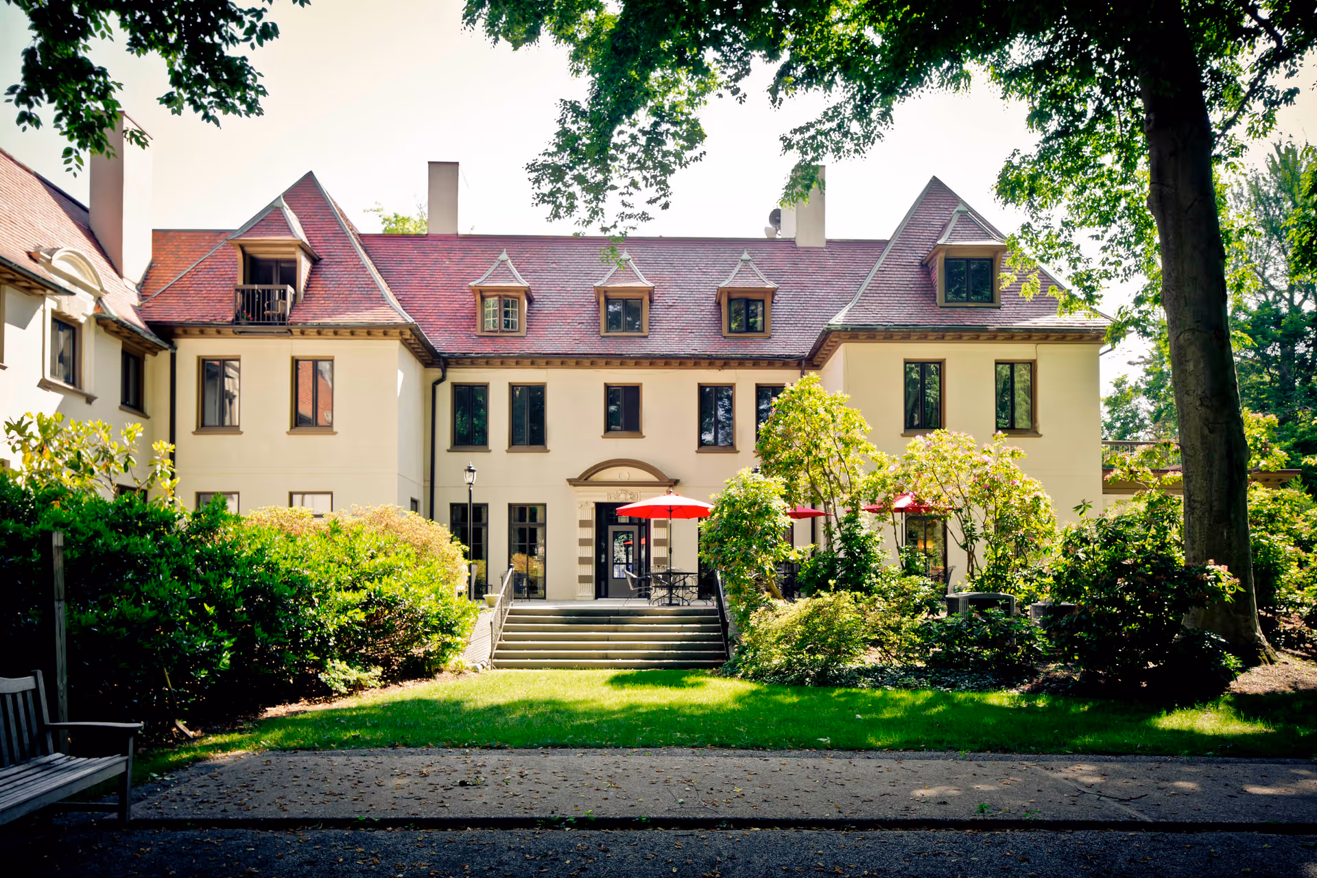 Front exterior of a large cream-colored mansion with a red roof, steps leading to a patio with red umbrellas, and surrounding trees and shrubs.