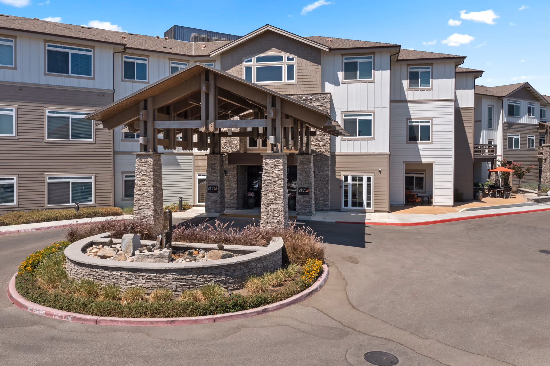 Front exterior view of a multi-story senior living facility with a covered entrance supported by stone pillars. There is a circular stone planter with landscaping and a small water feature in front of the entrance. The building has multiple windows and a mix of beige and white siding under a clear blue sky.