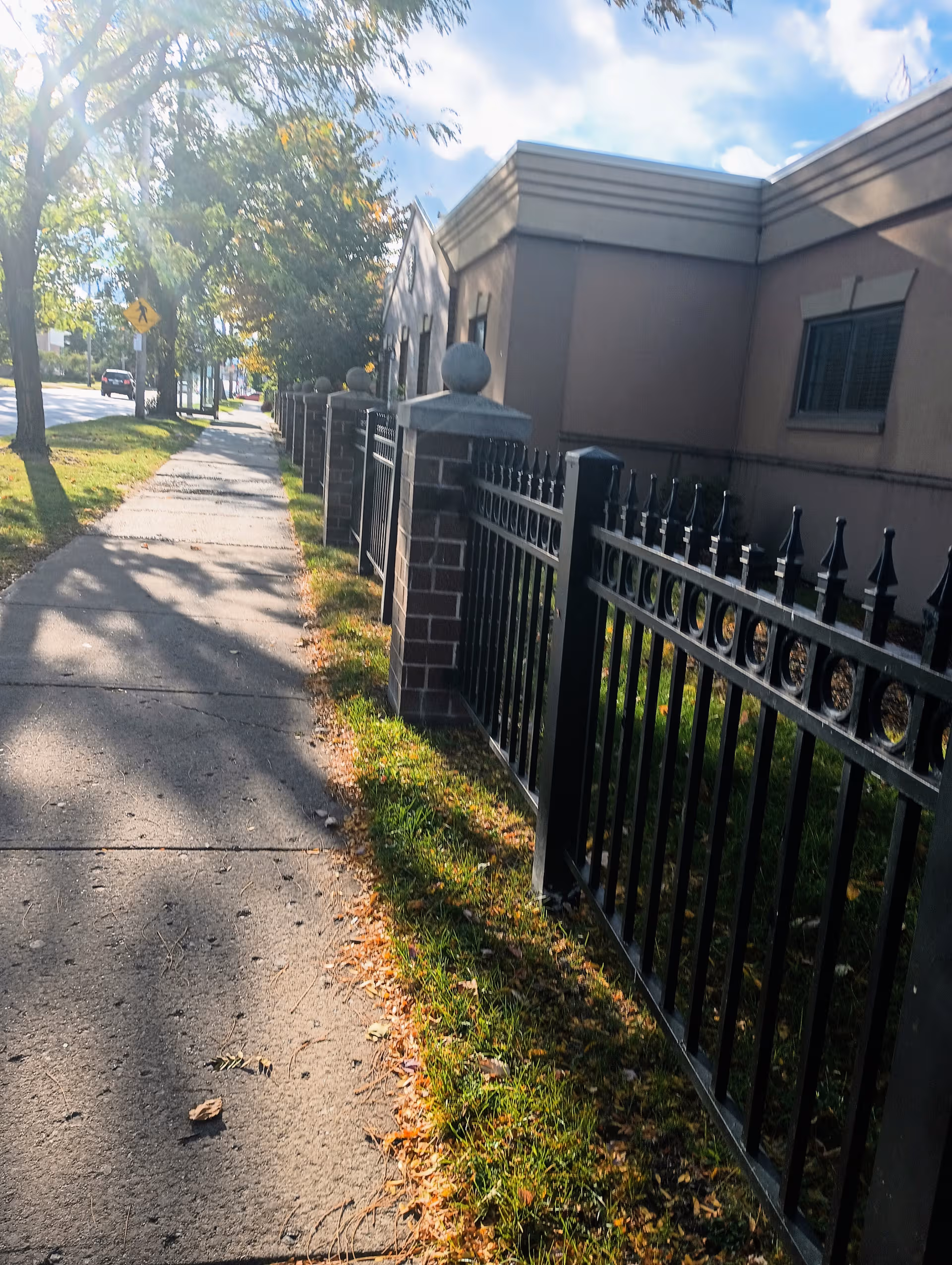 Sidewalk beside a beige facility with black wrought-iron fence and trees along the path.